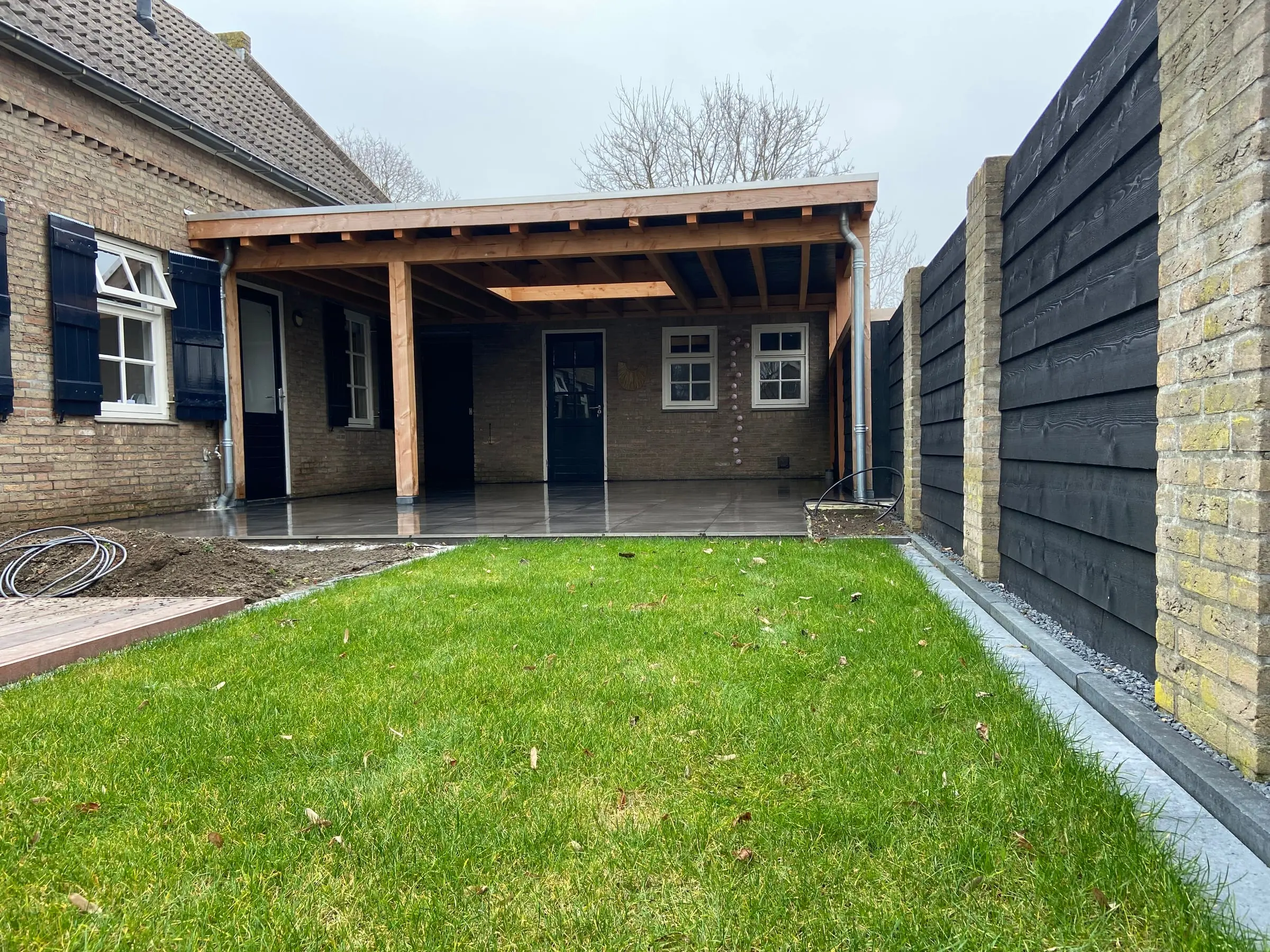 Grote houten overkapping over het terras, tegen een woning met blauwe luiken